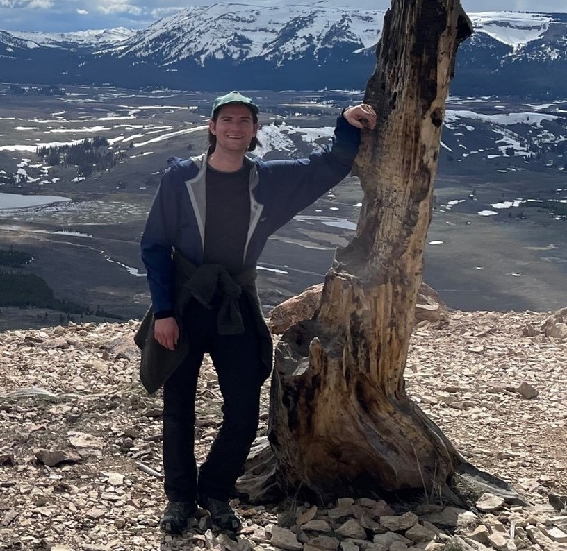 Alexander Allison leaning on a dead tree with mountains in the distance
