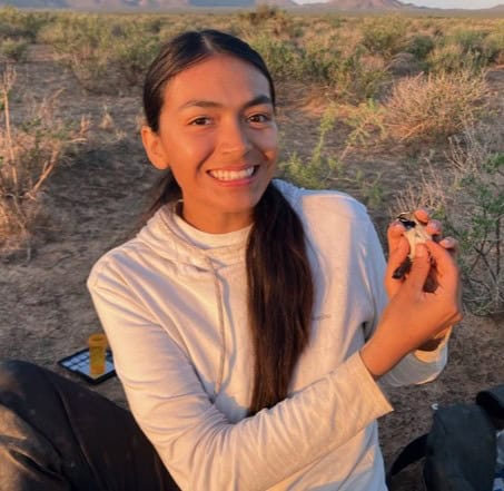 Amyliah Pina in the desert holding a small bird in her hands. A low sun emits golden light and casts shadows on the mountains behind her.