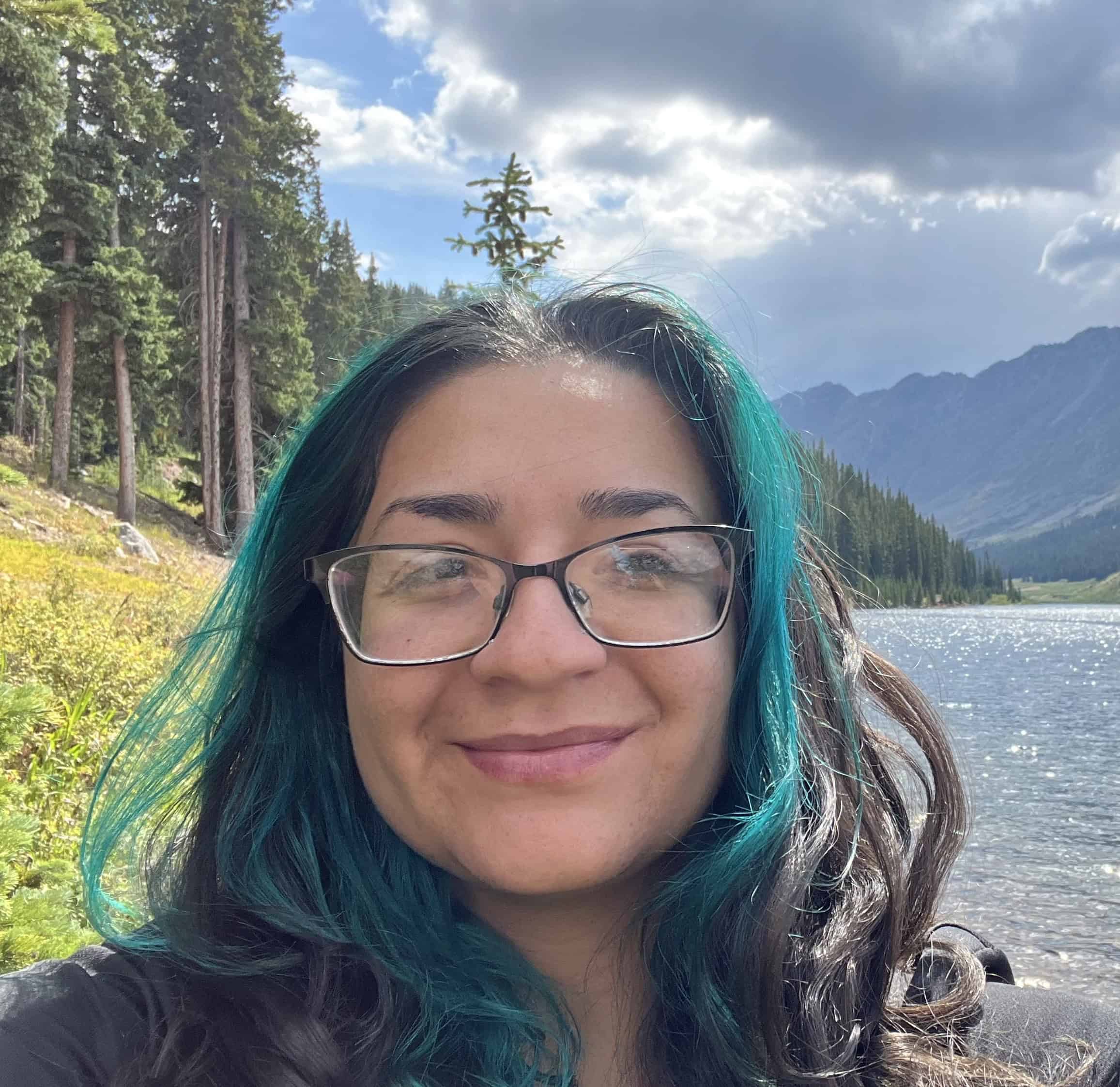 Brianna Perez smiles at the camera. Behind her are a grassy hill and trees to her right and a fresh body of water and mountains to her left.