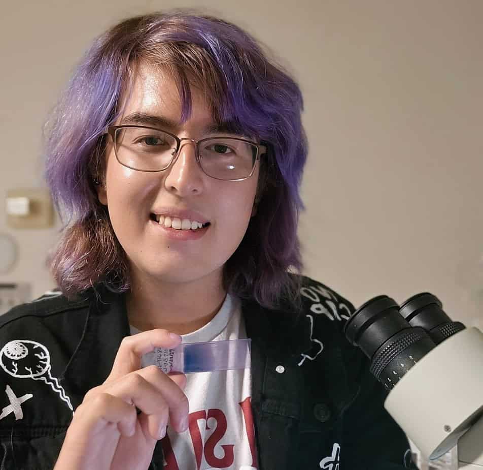 Edward Ortiz is holding a purple-stained slide, which matches the purple in his hair, and is smiling into the camera. The eye pieces of a camera peek through from the bottom right corner.