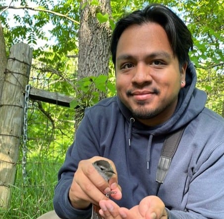 Eriberto Osorio smiling and holding a small gray bird in his hand. He is wearing a navy sweatshirt.