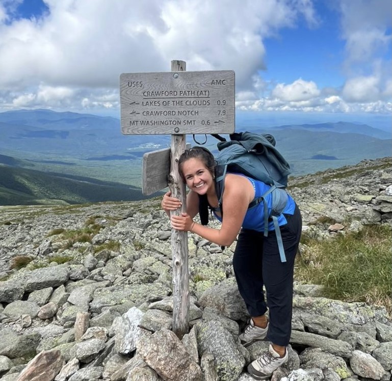 Photo of Fatima Quiroz holding a sign that reads "Crawford Path (AT)" with arrows pointing left towards "Lake of the Clouds 0.9" and "Crawford Notch 7.9" and an arrow pointing right towards "MT Washington SMT 0.6."