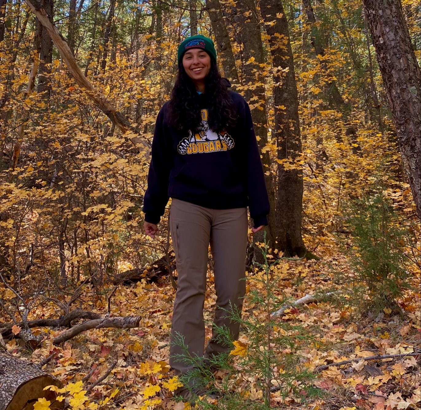 Grace Salmon standing in front of trees with golden leaves. She is wearing a beanie, a black sweatshirt with "Cougars" written on it, and brown pants.