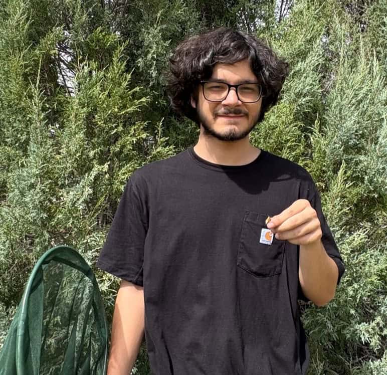 Jeremiah Fierro is standing before some trees in a black shirt and is holding a green net in one hand.