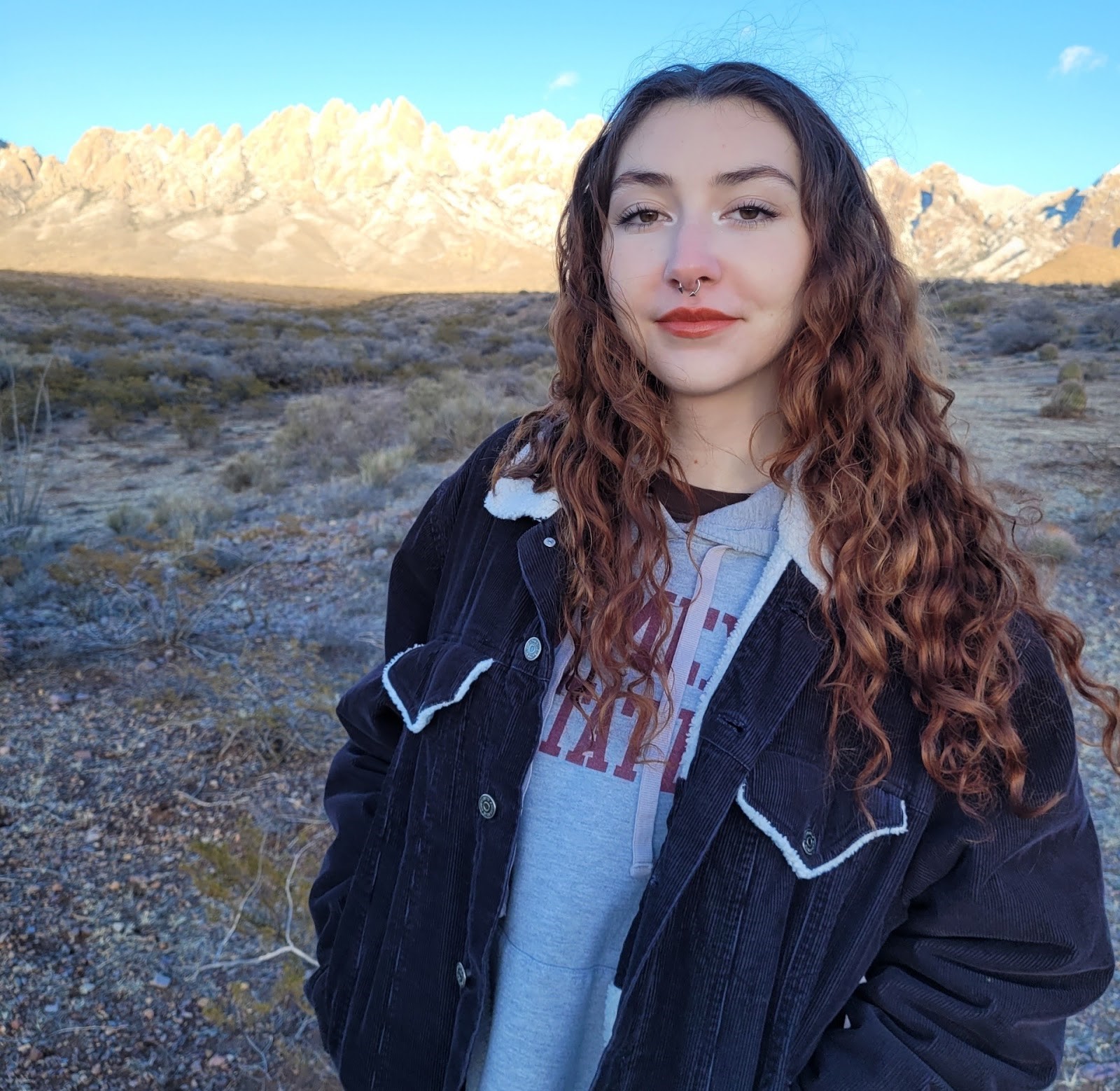 Julianna Diaz standing before the snow-covered Organ Mountains. She has brown curly hair and is wearing a black jacket over a gray New Mexico State sweatshirt.