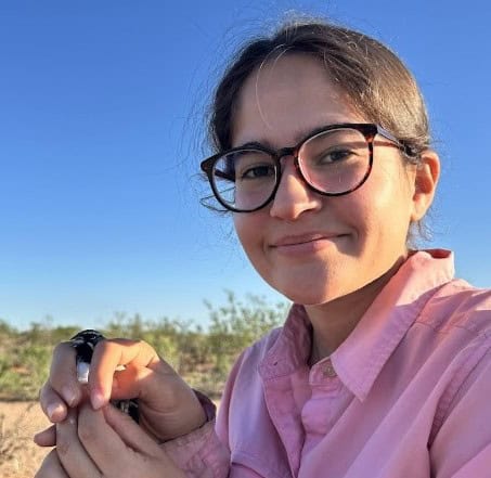 Juliemar crouches in the desert holding a bird in a pink shirt and black-framed glasses.
