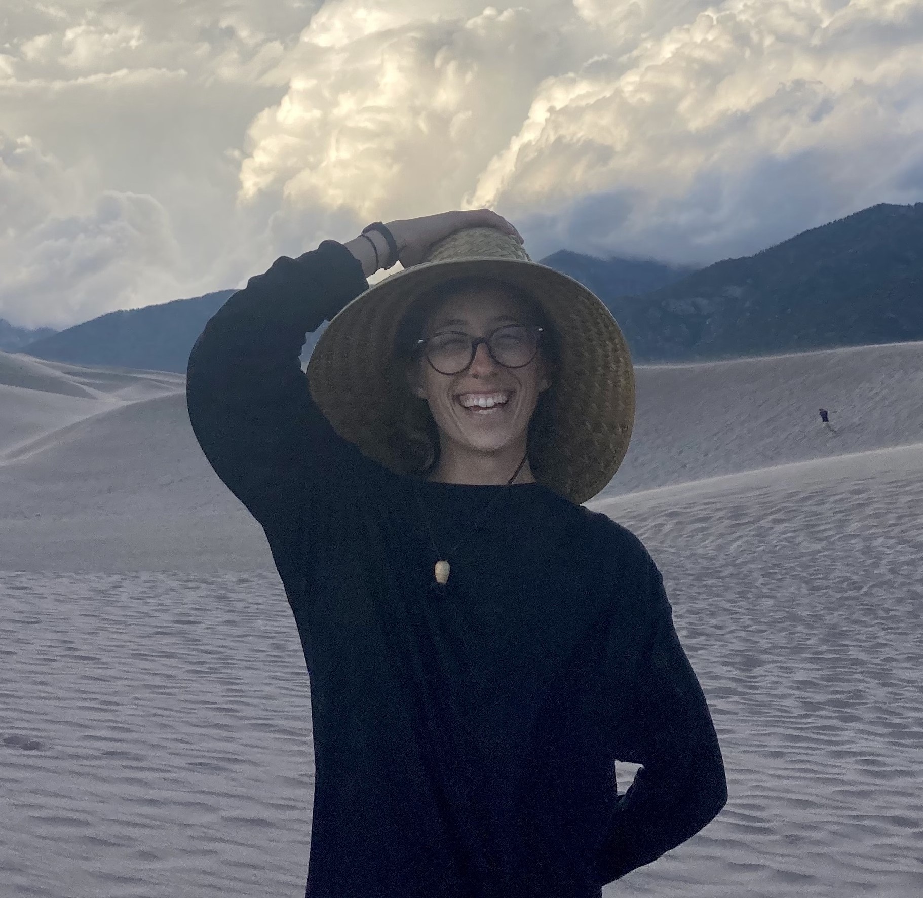 Photo of Kelley Boland smiling and holding a hat onto her head in front of rolling hills of sand, some mountains, and a sky hidden by clouds.