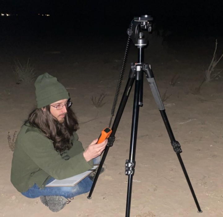 Photo of Thomas Huycke sitting cross-legged in the desert at night, looking at an orange reader hooked up to a tripod
